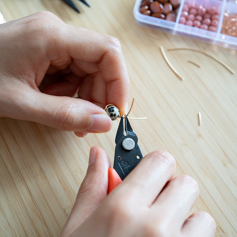 Persona usando pinzas de bisuteria para trabajar una cuenta sobre una superficie de madera.