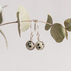 Par de aretes plateados con piedras moteadas colgando de una rama con hojas.