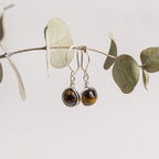 Aretes plateados con piedras ojo de tigre sobre rama con hojas.