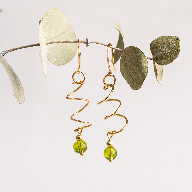 Aretes dorados en espiral con cuentas verdes piedranatural peridoto, piedra de agosto sobre rama con hojas, joyería artesanal.