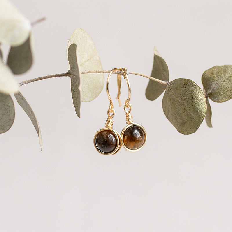 Aretes dorados con piedras cafes sobre rama de eucalipto.