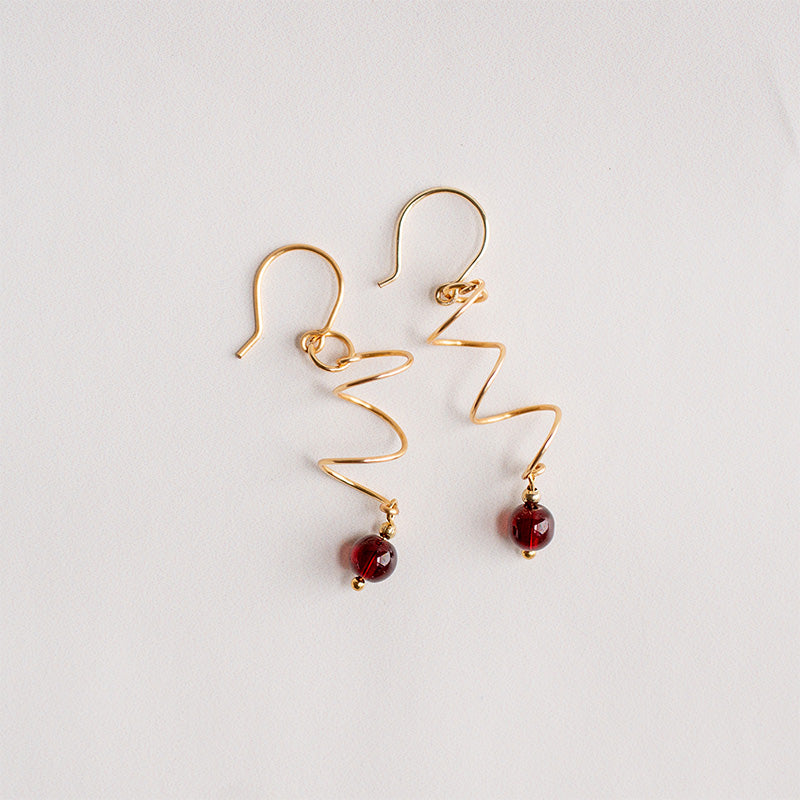Aretes dorados en espiral con cuentas rojas garnet piedra natural sobre fondo blanco, joyería artesanal.