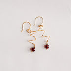 Aretes dorados en espiral con cuentas rojas garnet piedra natural sobre fondo blanco, joyería artesanal.