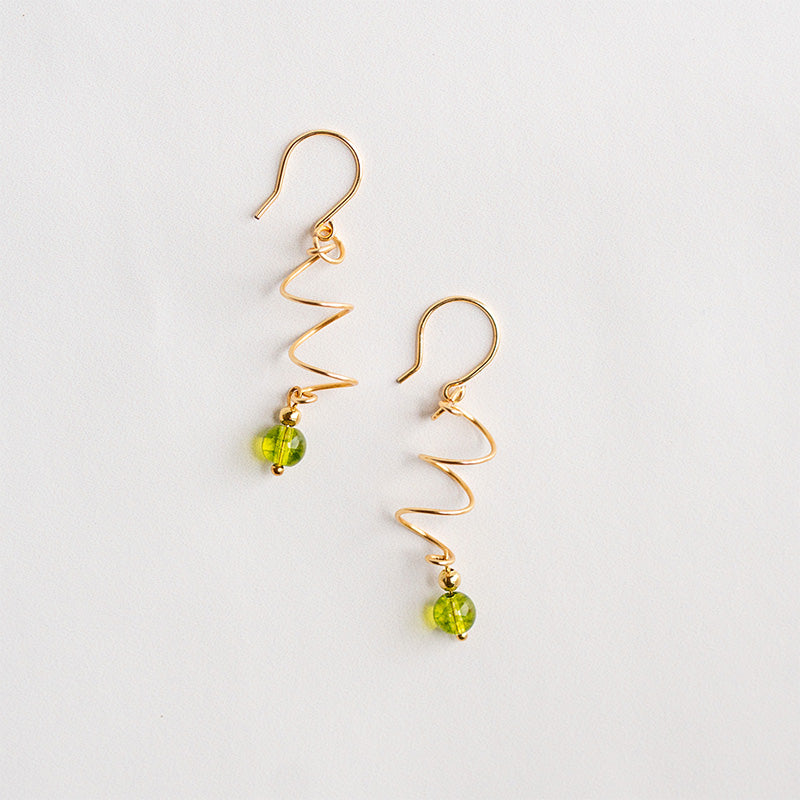 Aretes dorados en espiral con piedra natural peridoto verde sobre rama con hojas, joyería artesanal.
