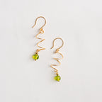Aretes dorados en espiral con piedra natural peridoto verde sobre rama con hojas, joyería artesanal.