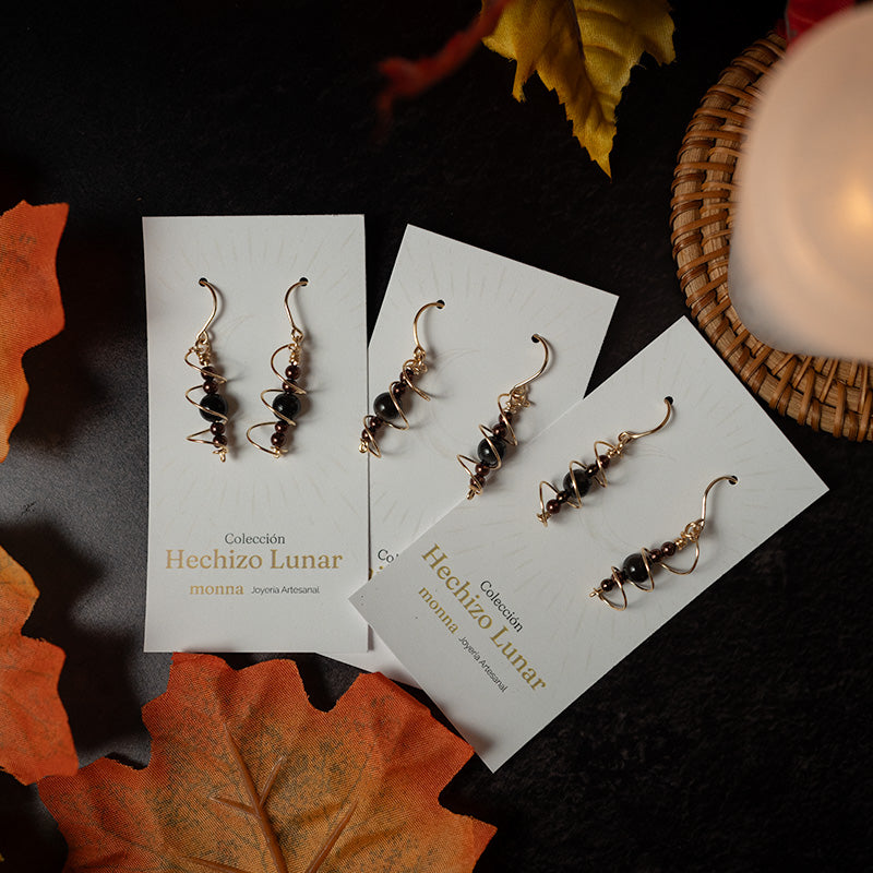Par de aretes dorados y negros sobre tarjetas de Hechizo Lunar, con hojas otoñales y una vela al fondo.
