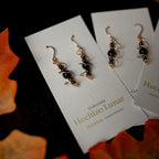 Par de aretes dorados y negros en espiral sobre una tarjeta de “Hechizo Lunar”, con fondo oscuro.