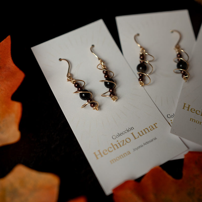 Par de aretes dorados y negros en espiral sobre una tarjeta de “Hechizo Lunar”, con fondo oscuro.