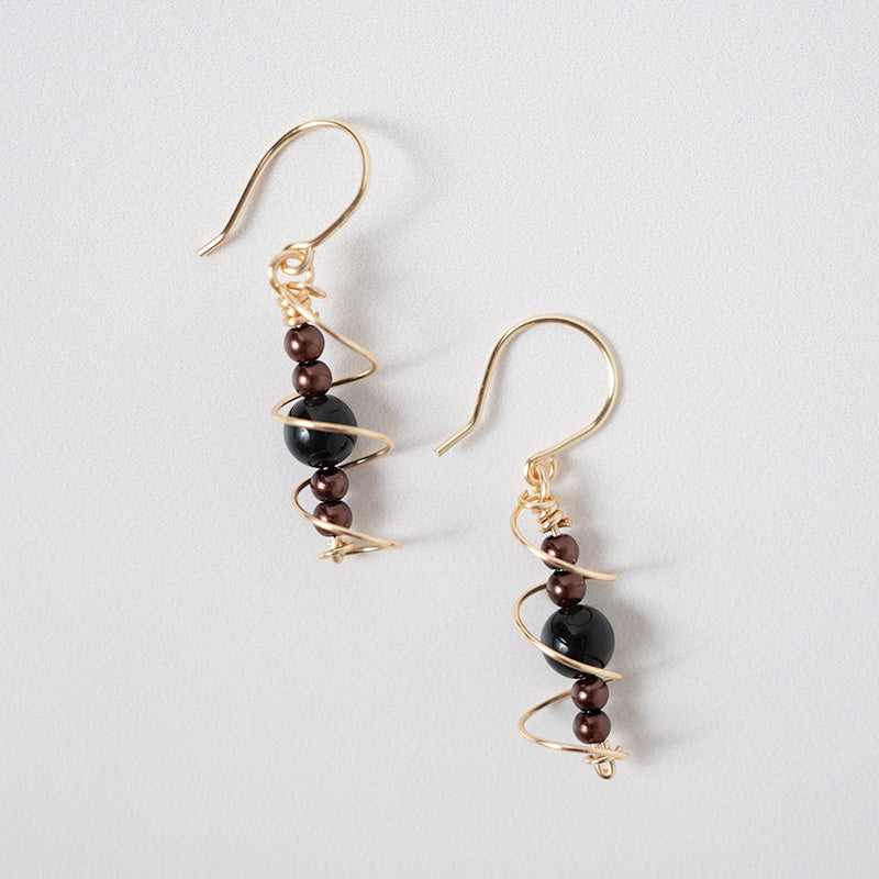 Par de aretes dorados en espiral con cuentas negras y marrones sobre un fondo claro.