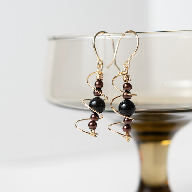 Par de aretes dorados y negros con cuentas sobre un soporte, con fondo blanco