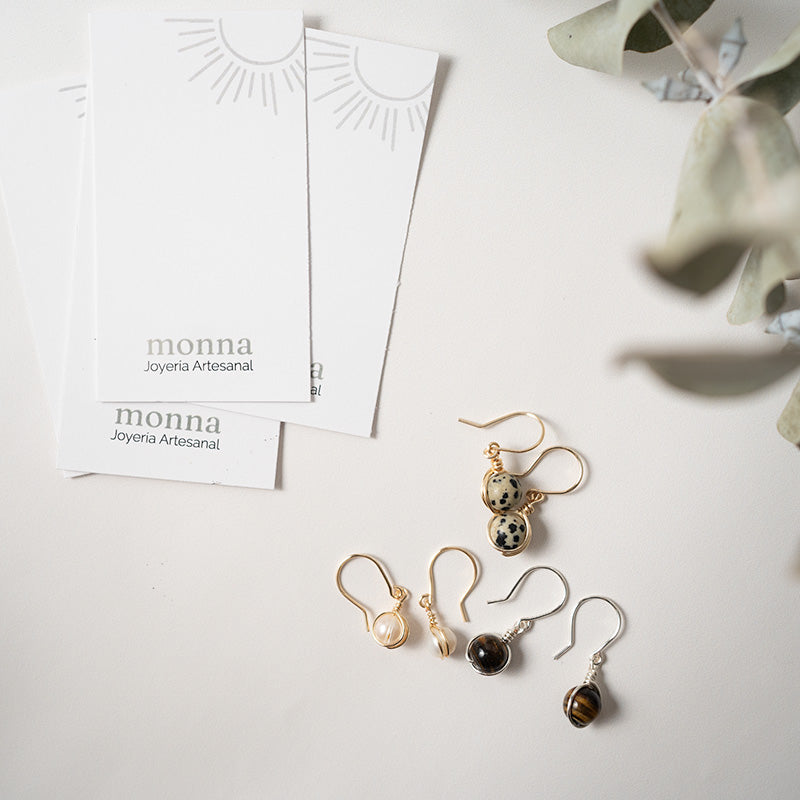 Juego de aretes con tarjetas de “monna Joyería Artesanal” sobre una superficie blanca.
